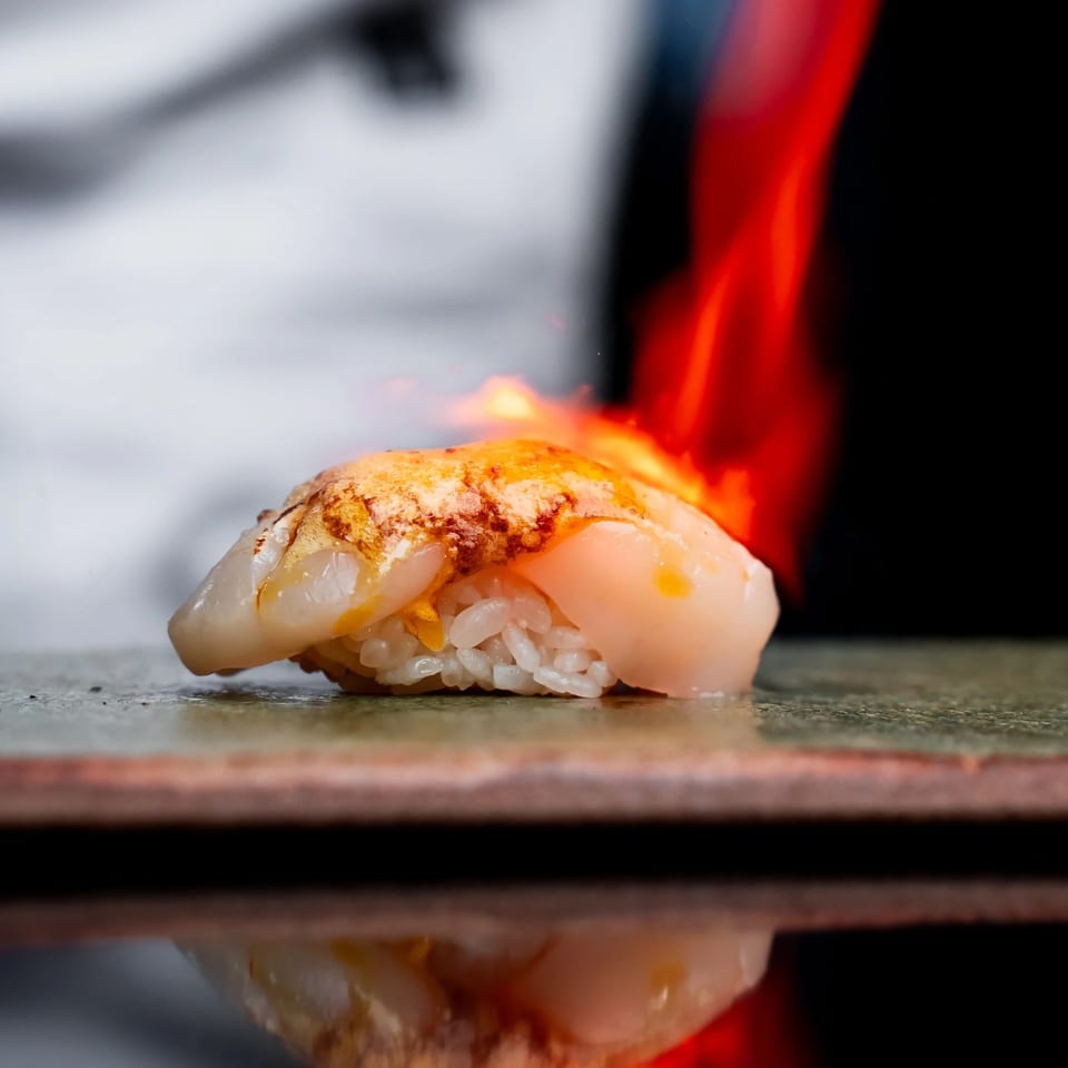 Close-up of glazed sushi nigiri with white fish over rice, reflected on a counter against dramatic red lighting.