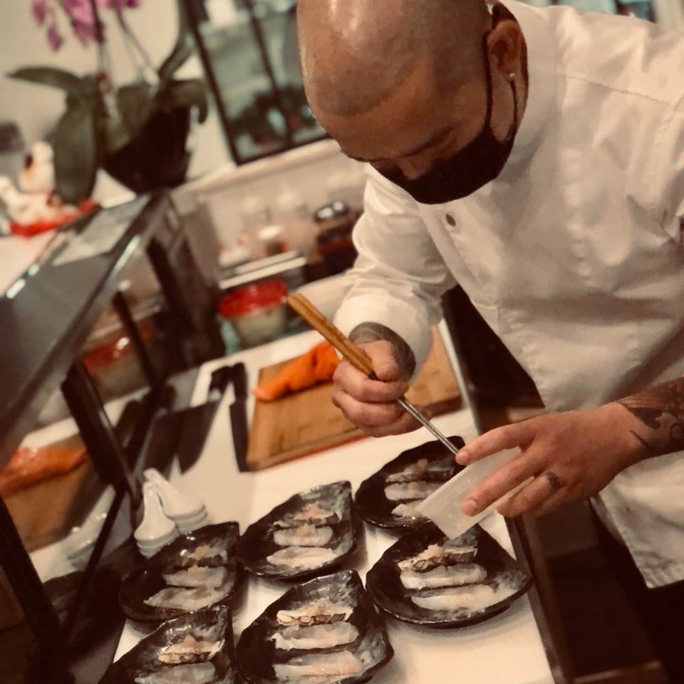 Chef in a white coat uses chopsticks to arrange sushi rolls on a platter in a warmly lit restaurant.