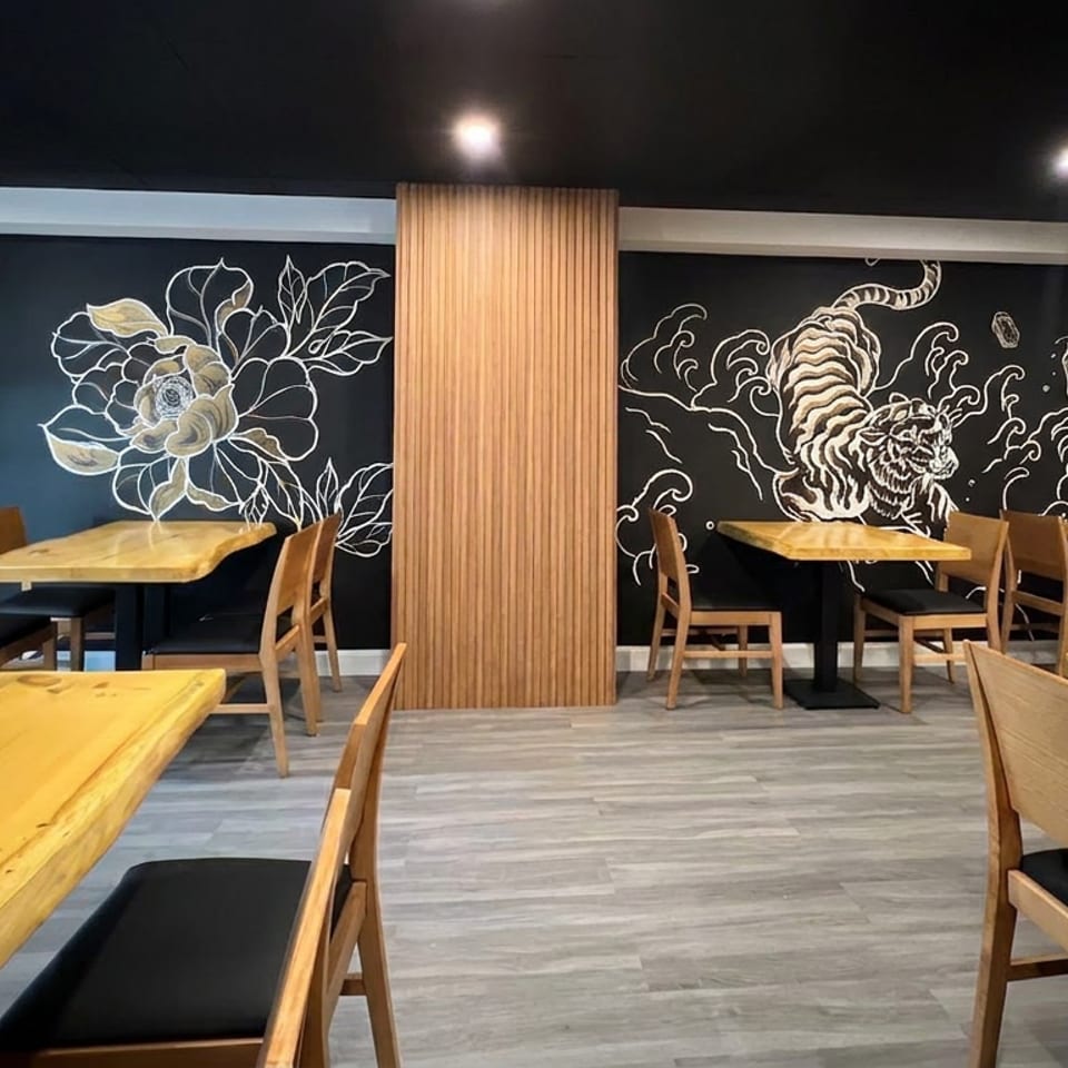 Modern restaurant dining room with wooden tables, chairs, black ceilings, and large floral and ocean-themed wall murals.
