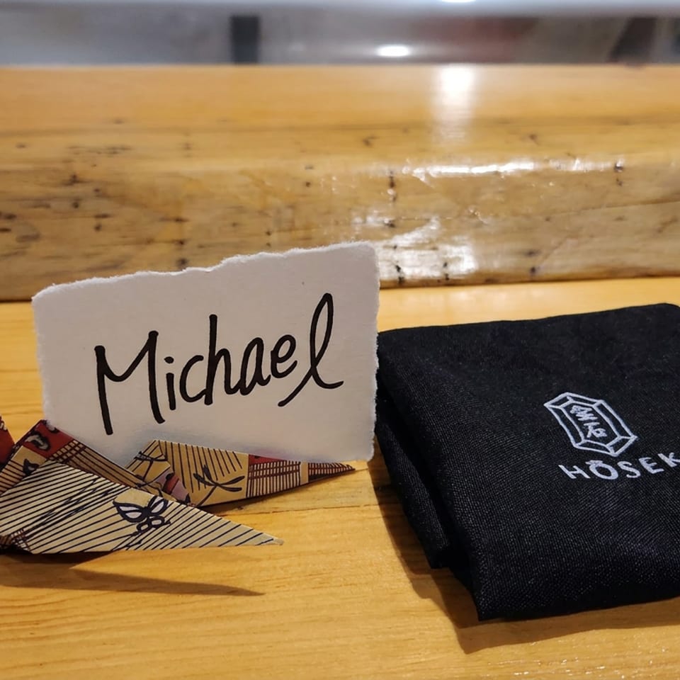 Handwritten “Michael” place card beside a patterned origami crane and folded black HOSEKI napkin on a wooden restaurant table.
