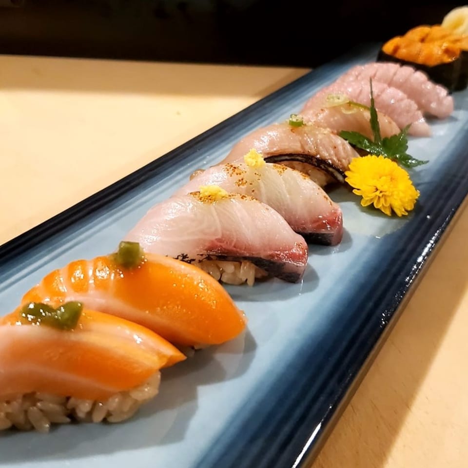 Assorted sushi nigiri arranged on a long blue platter, garnished with wasabi, ginger, and a yellow flower.