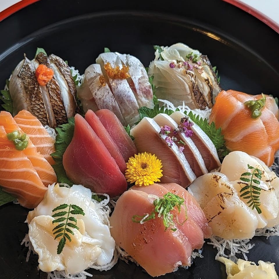 Assorted sashimi, including salmon, tuna, scallops, and seared fish, arranged on shredded daikon with flowers and herbs.