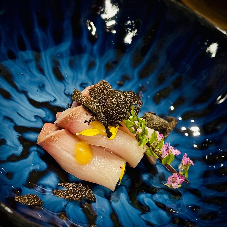 Delicate seafood dish topped with black truffle slices, yellow petals, greens, and pink flowers on a glossy blue plate.